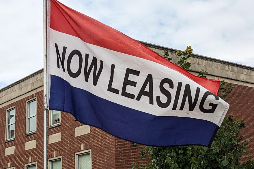 A red, white and blue flag waves in front of a brick building with NOW LEASING written in black letters.