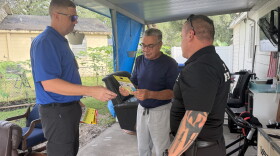 Three men stand in a half-circle talking to each other, with the one in the middle reading a yellow flyer. They are standing under a covered area outside of a house, with grass, another house and household items in the background.