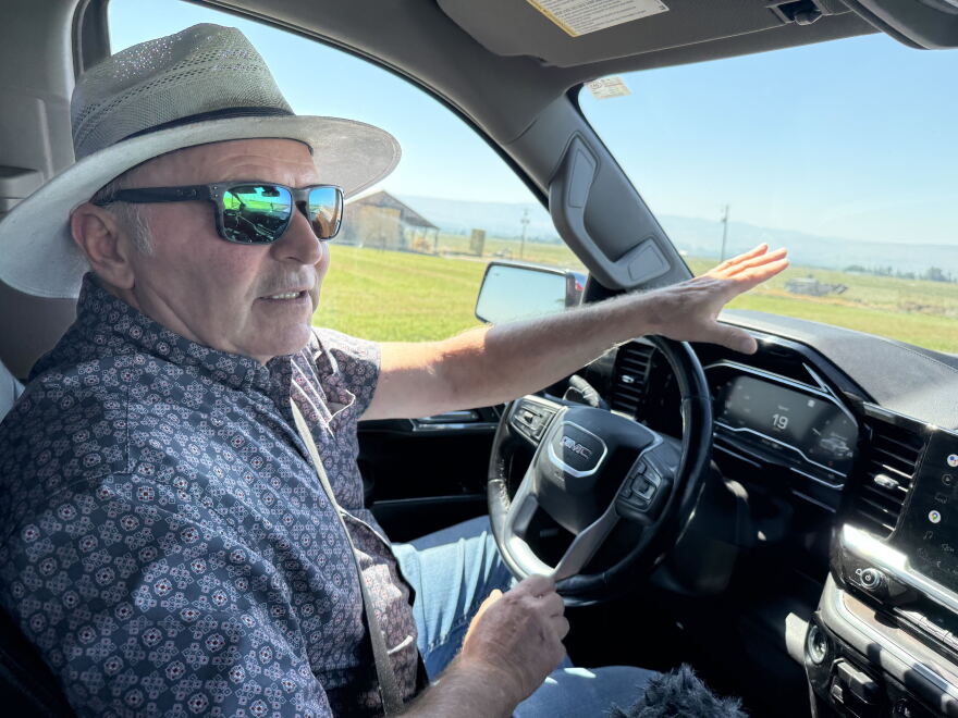 A man inside the cab of a pickup truck driving around a farm pauses to talk, gesturing to the fields outside his windshield.