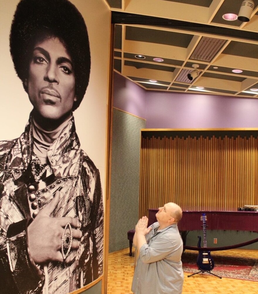  Phil Harrell kneels in a praying position and looks up at a huge photo of Prince. The room has high acoustic ceilings, wood floors and a piano.