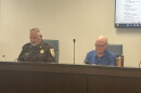 An image of a man in sheriff's department uniform and another man sitting on a dais in a meeting of the McLean County Emergency Telephone System Board in McLean County.