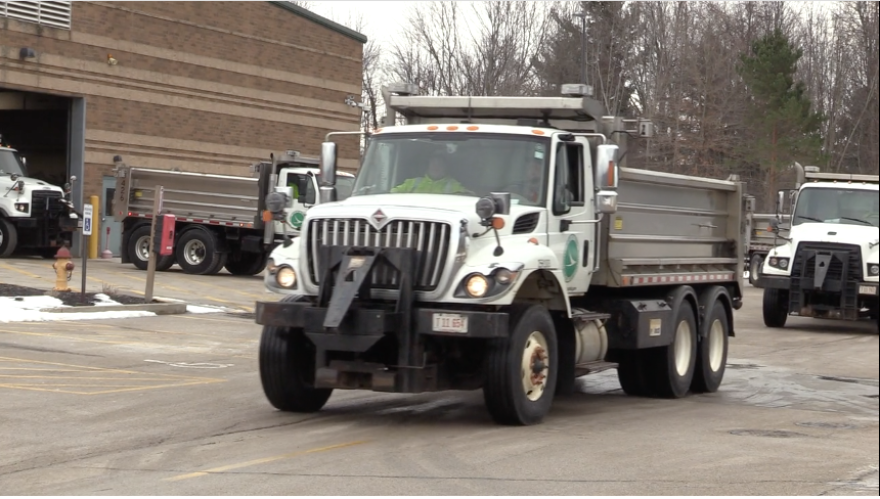 Two people sit in the cab of a large truck in the middle of a parking lot. The truck appears to be moving forward. Three more trucks are in the background.