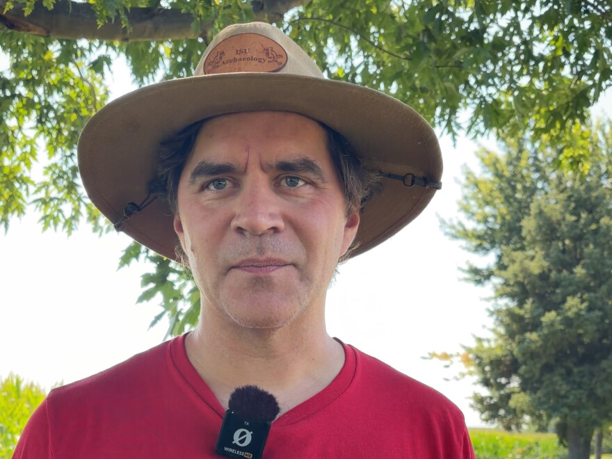 a man wearing a red t-shirt and brown sunhat stands in front of green trees