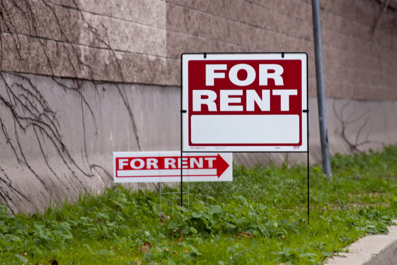 two red and white for rent signs are posted in a grassy strip next to a grey brick wall.