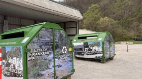 Collection bins at the City of Frankfort Recycling Center. The bins are about 5 feet tall with a footprint of about 4x7 feet. They're green, dumpster-shaped, with slits to receive recycling on both of the wider sides. Two of them are in the frame, sitting outside of an open covered space protecting additional bins from the weather.