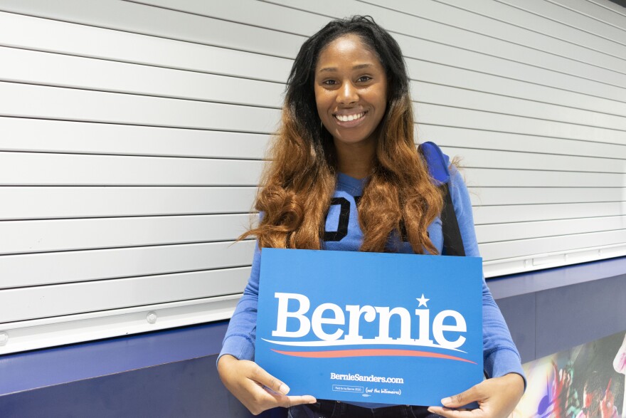 A woman smiles for a photo and holds a blue sign that says Bernie on it.