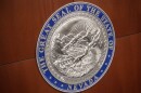 A close-up of the seal of Nevada. There are two borders with words. The outer border says, “The Great Seal of the State of Nevada.” The inner border says, “All for our Country.” The center is artwork that includes a quartz mill, train, mountains and more.