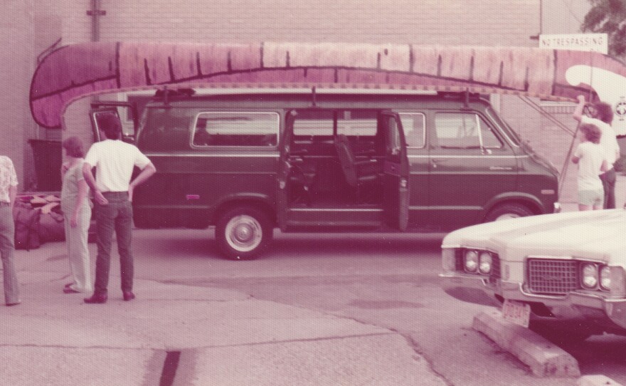 A long voyageur style canoe sits atop a van in 1976.