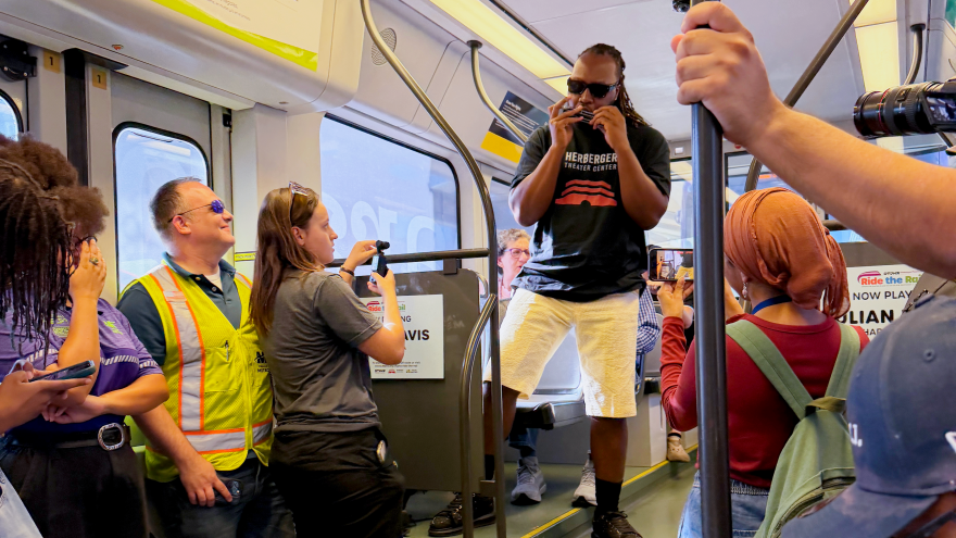 Man in black shirt and sunglasses plays the harmonica on a light rail train