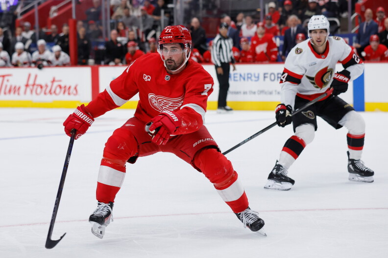 Detroit Red Wing Dylan Larkin in a red jersey skates with the puck, pursued by Ottawa Senators player Dylan Cozens in a white jersey.