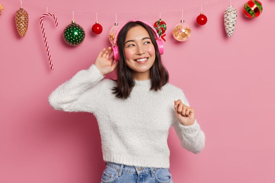A woman smiles as she listens to music on pink headphones in front of Christmas decorations
