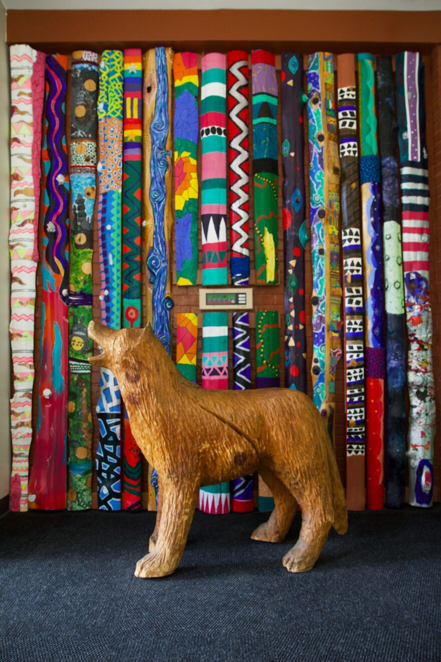 A wooden carved wolf stands in front of colorfully-painted slabs of wood lined up against a wall.