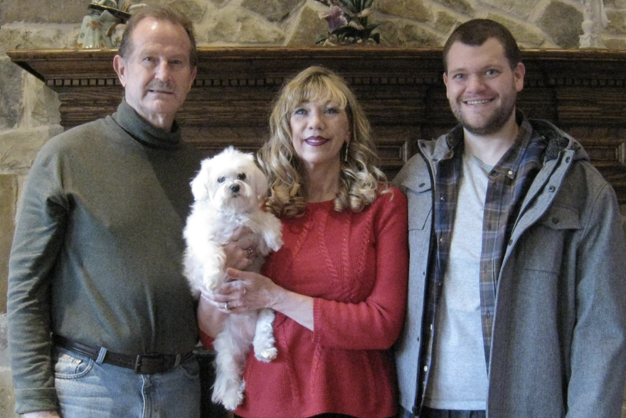 The family stands in front of a fireplace. Frank has brown hair and a mustache and is wearing a green turtle-neck shirt. Jackie is a blond woman with a red shirt and is holding the family's small, white dog. Bryce has brown hair with fivo-o-clock shadow and is wearing a white tee shirt with a flannel shirt and a gray jacket over it. 