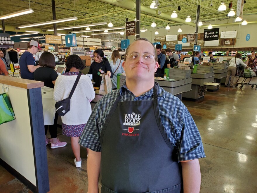 a man wearing glasses, a short-sleeve button-up plaid shirt and an apron with the whole foods logo smiles in front of a row of supermarket registers