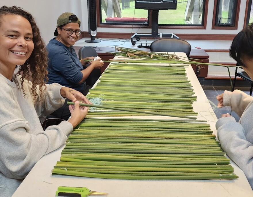 From L to R: Volunteers Alayne Elizabeth Grigsby, Priya Bhattacharjee, and Amanda Cermelita Oguara prepare to weave tule into mats for serving food on.