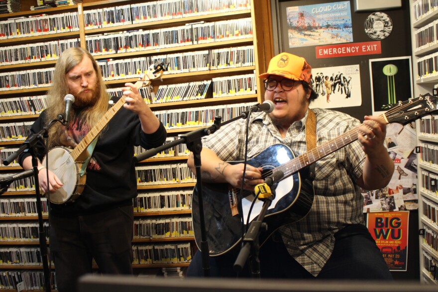 A person playing banjo stands to the left of a seated person playing guitar in a radio studio. Behind them are walls covered with shelves of CDs and a door that says Emergency Exit that has band posters on it.