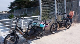 Two large e-bikes are parked against a fence with helmets hanging from them. One has a long seat for two kids and the other has a seat with a harness for one child. 