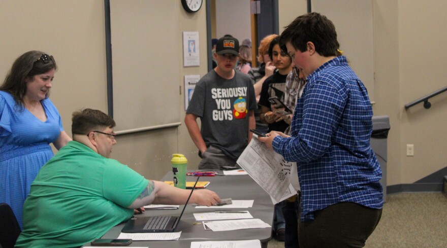 Several people stand at a table, where a person seated with a laptop assists another person holding papers. Others wait in line behind them in a room with beige walls.