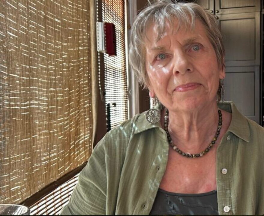 Older woman with grey hair wearing green button down shirt sits inside near a window 