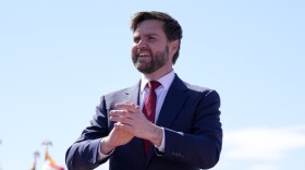 a man in a suit stands smiling and clapping while an american flag flies behind him