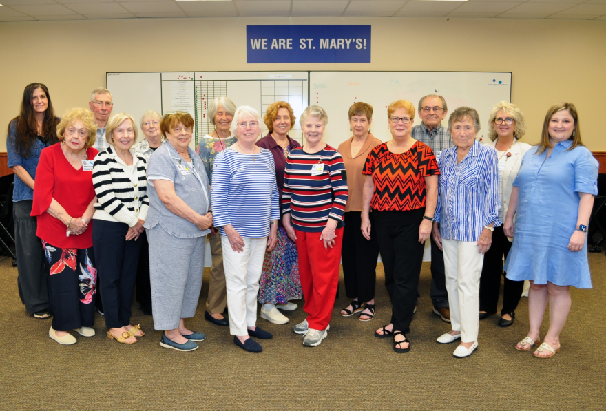 A group of hospital volunteers and staff pose together in a room beneath a sign reading, “We are St. Mary’s!”