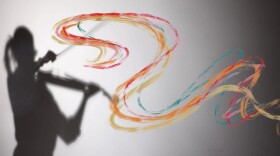 The silhouette of a violinist is cast upon the left side of a screen, created by a single spotlight. Colorful painted streaks of yellow, pink, orange, red blue are twisting and weaving across the image, originating from the violin's shadow and traveling to the right.