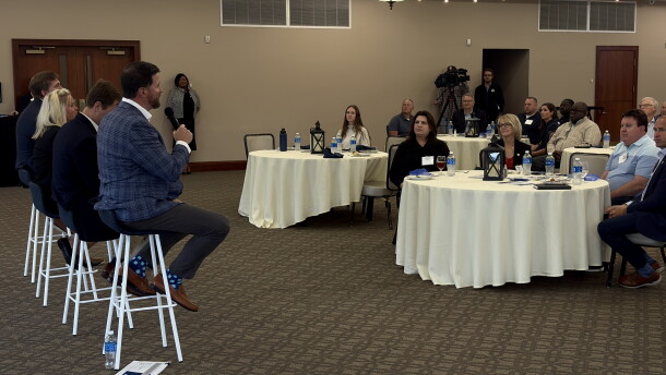 Four individuals sit on stools in front of people sitting at several round tables with white tablecloths. The person closest to the camera, on a stool,  is holding a microphone. 