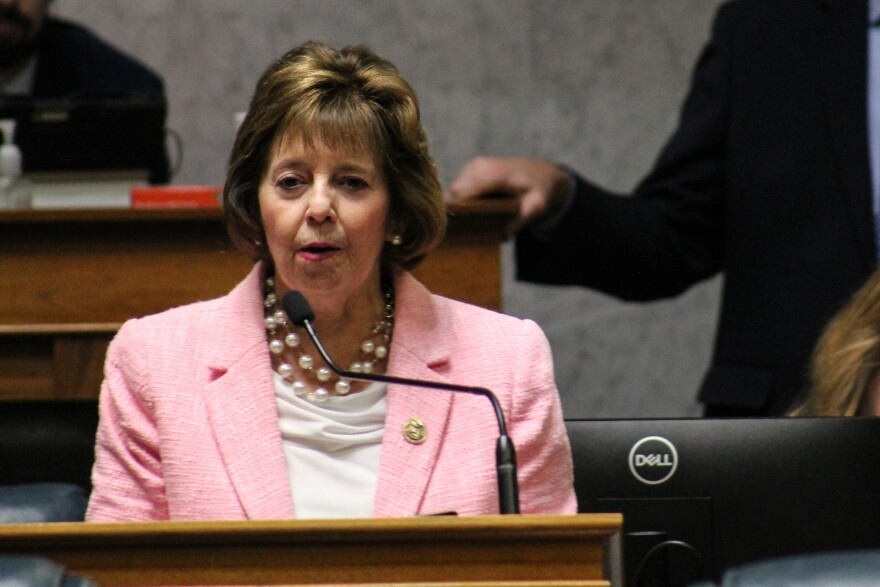 Senator Linda Rogers stands at a podium in Indiana Senate. She is a White woman with brown hair and wears a pink suit.