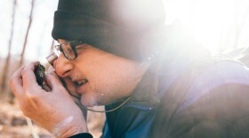 A white man wearing a black beanie looks at moss through a hand lens.
