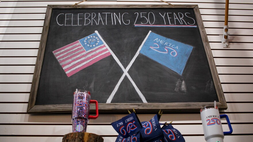 Two flags are drawn on a chalkboard: An American flag with "250" and circle of stars where the 50 stars usually are, and another flag that reads "America 250." Smaller America 250 flags and two America 250 cups sit below the board. 