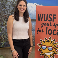 A woman in a white shirt and dark pants stands next to a sign reading "WUSF" that has a large graphic of a smiling sun. 