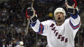 Chris Simon holds up both arms shouting with a hockey stick in his right hand and wearing full white hockey uniform with the word "Rangers" printed on the jersey. Stadium seats are in the background.