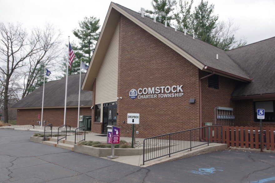 Gray clouds hang over a long brick building, the center of which sticks out with its almost tent-like peak. "Comstock Charter Township" is written on the peaked front of the building in silver letters, along with the blue and gray logo. Three flags, one of which is clearly the American flag, sit on poles to the right of the image. 