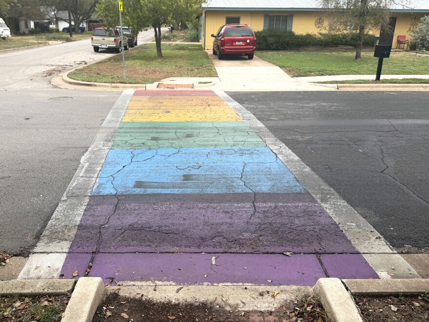 A rainbow crosswalk with alternating colors of purple, blue, green, yellow and red. Thick white lines run on the outsides of the crosswalk. It is partially faded from being driven over. In the background is a maroon minivan parked in the driveway of a yellow house with green trim. 