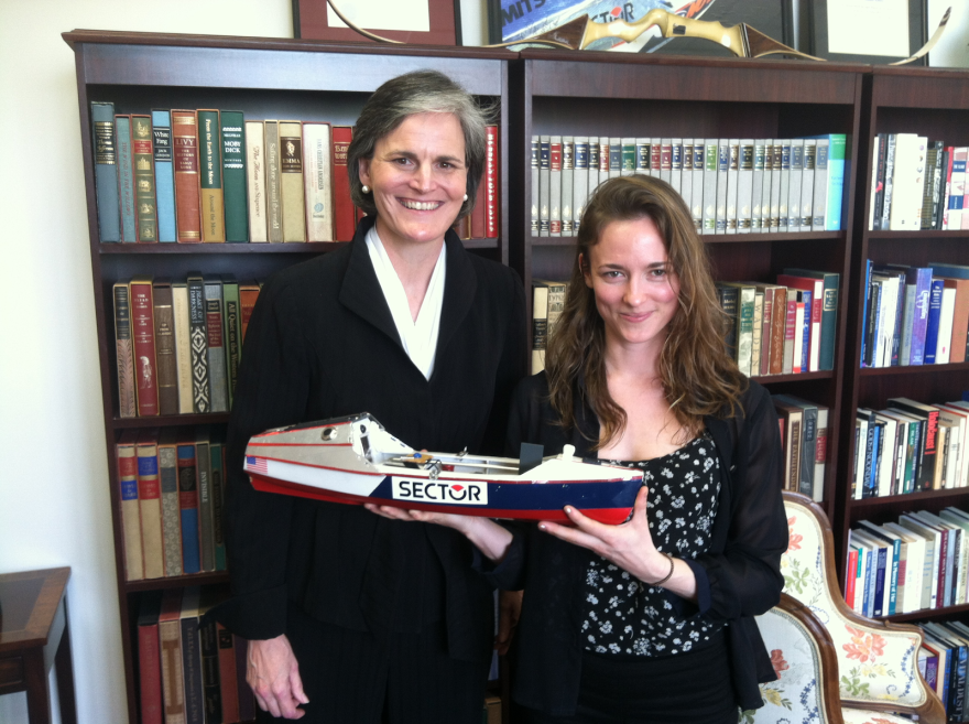 Toni Murden McClure, a middle aged white woman with brown and grey hair standing next to Dawn Landes, a white woman with brown hair. Both are smiling and holding a boat figurine