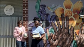 Two women stand outside The Little Resource Center in Carbondale, holding books and a donation jar in front of a colorful mural.