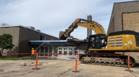 An excavator demolishes part of the front of a building while a man wearing orange sprays the site with a hose.
