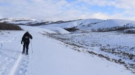 A person in cold-weather gear hikes with a pole through a snow-covered landscape of hills