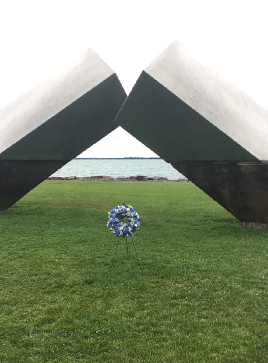 A wreath of purple and white flowers sits under a memorial on a green lawn.