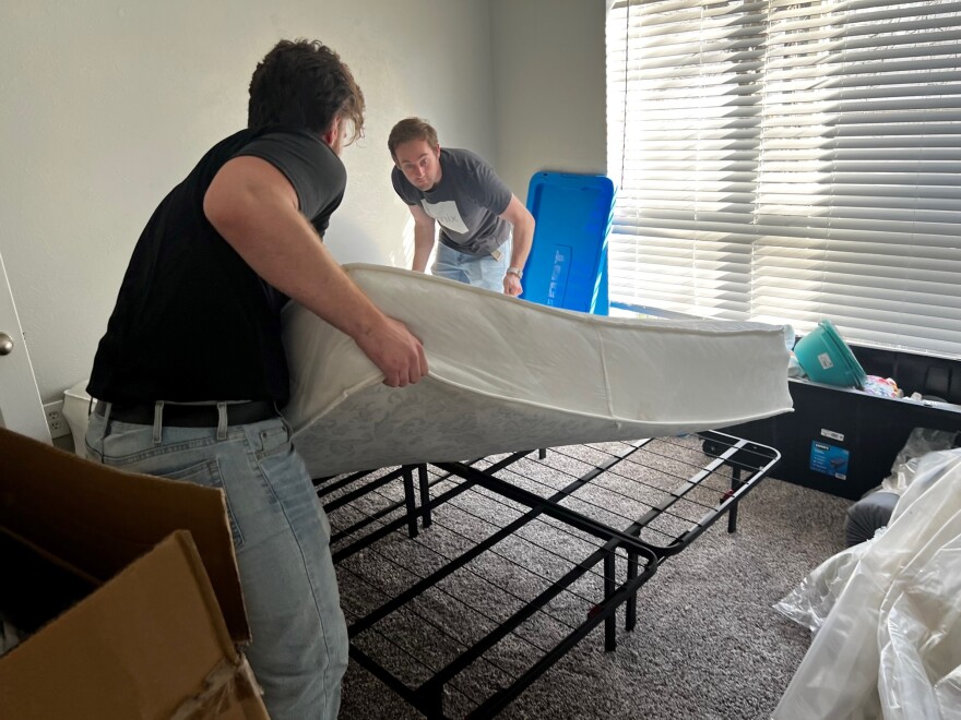 Two young men place a mattress on top of a bed frame inside an apartment.