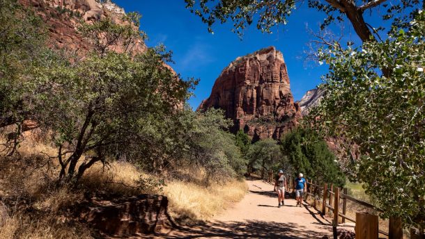 Two people walk along a trail with beautiful red rock formations in the background.