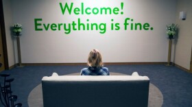 A woman sitting on a couch looking at a wall with green lettering that reads "Welcome! Everything is fine."
