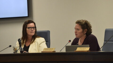 Two women sit at a desk with microphones. 