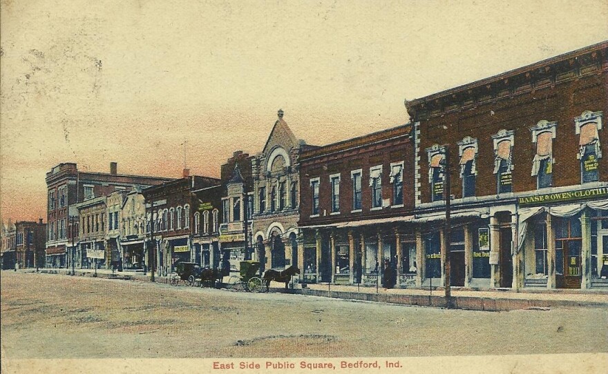 A colorized postcard showing a street view of the East Side Public Square in Bedford, Indiana during the early 1900s, featuring multi-story brick storefronts, horse-drawn carriages along the unpaved street, and text at the bottom reading "East Side Public Square, Bedford, Ind."