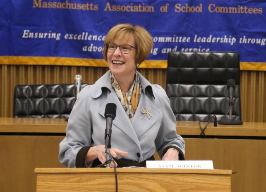 A white woman with glasses stands behind a podium smiling