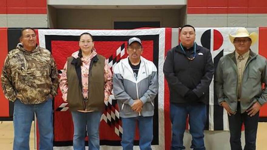 Five adults stand in front of a large quilt and the Northern Arapaho flag in a gym.