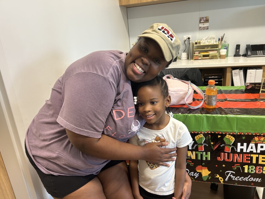 Sinquese Savage, a Black woman wearing a tan baseball cap and purple t-shirt, smiles at the camera while hugging her daughter, a little girl with her hair tied up with white beads at the ends of her braids. They stand in the entryway of a small daycare center that has a celebratory Juneteenth banner.