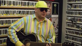 Man in bright yellow hat, brightly striped shirt, and sunglasses sitting on a stool playing guitar