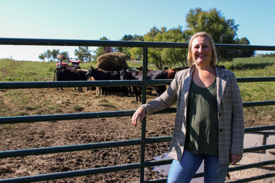 Jaqueline Witte stands in front of the cows eating hay. Her arm is propped on the gate, which separates them.