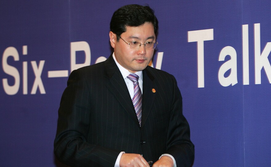 A man wearing a dark suit, tie and glasses holds a piece of paper while looking downward. Behind him is a blue background with white lettering, partially obscured, spelling out "Six-Party Talks."